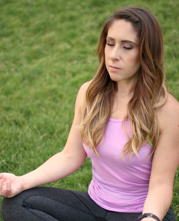 Photo of Andie outdoors meditating in the grass eyes closed and really relaxing in the moment and enjoying the benefits of nature. 