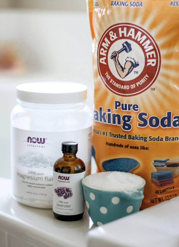 Photo of ingredients used in detox bath recipe magnesium flakes, lavender essential oil, Epsom salt, and baking soda. Sitting on the edge of a white tub. 