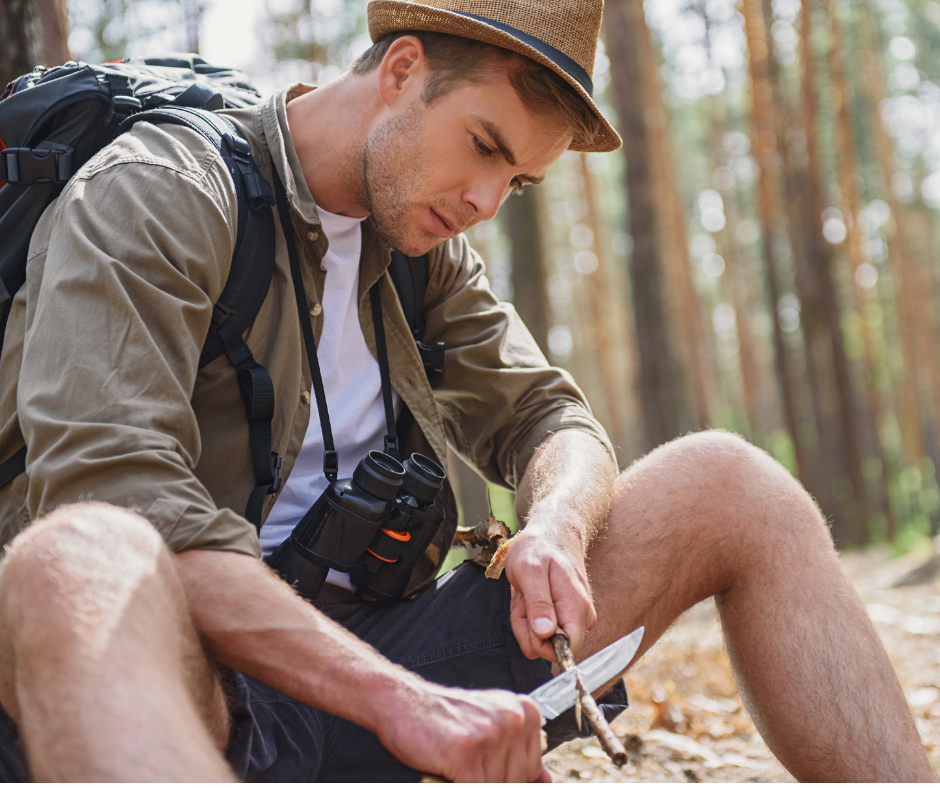 Photo of man in camping gear in the woods sitting on a log, whittling a stick with a sharp knife. Enjoying some time alone in the great outdoors. 